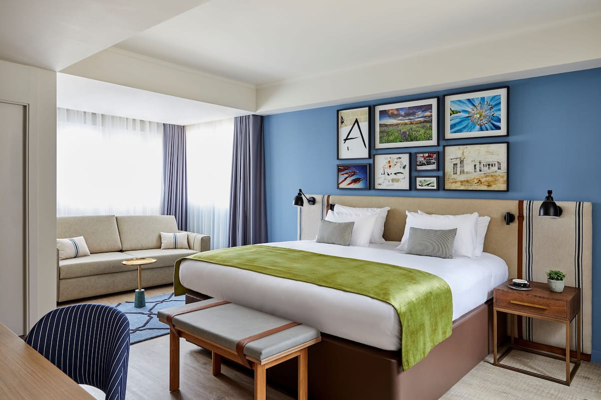 A hotel room with a large bed, a sofa, a desk, and a chair. There are framed pictures on the wall and a window with curtains.