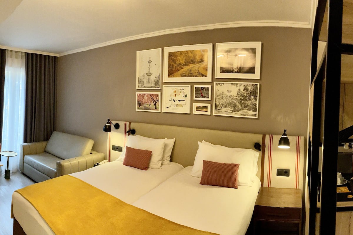 A hotel room with a large bed, a sofa, a desk with a chair, and a dining table. There are framed pictures on the wall and a coat rack with a hanging item.