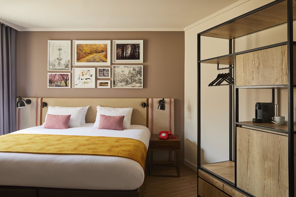 A hotel room with a large bed, a wooden wardrobe, a nightstand with a lamp, and a wall with framed pictures.
