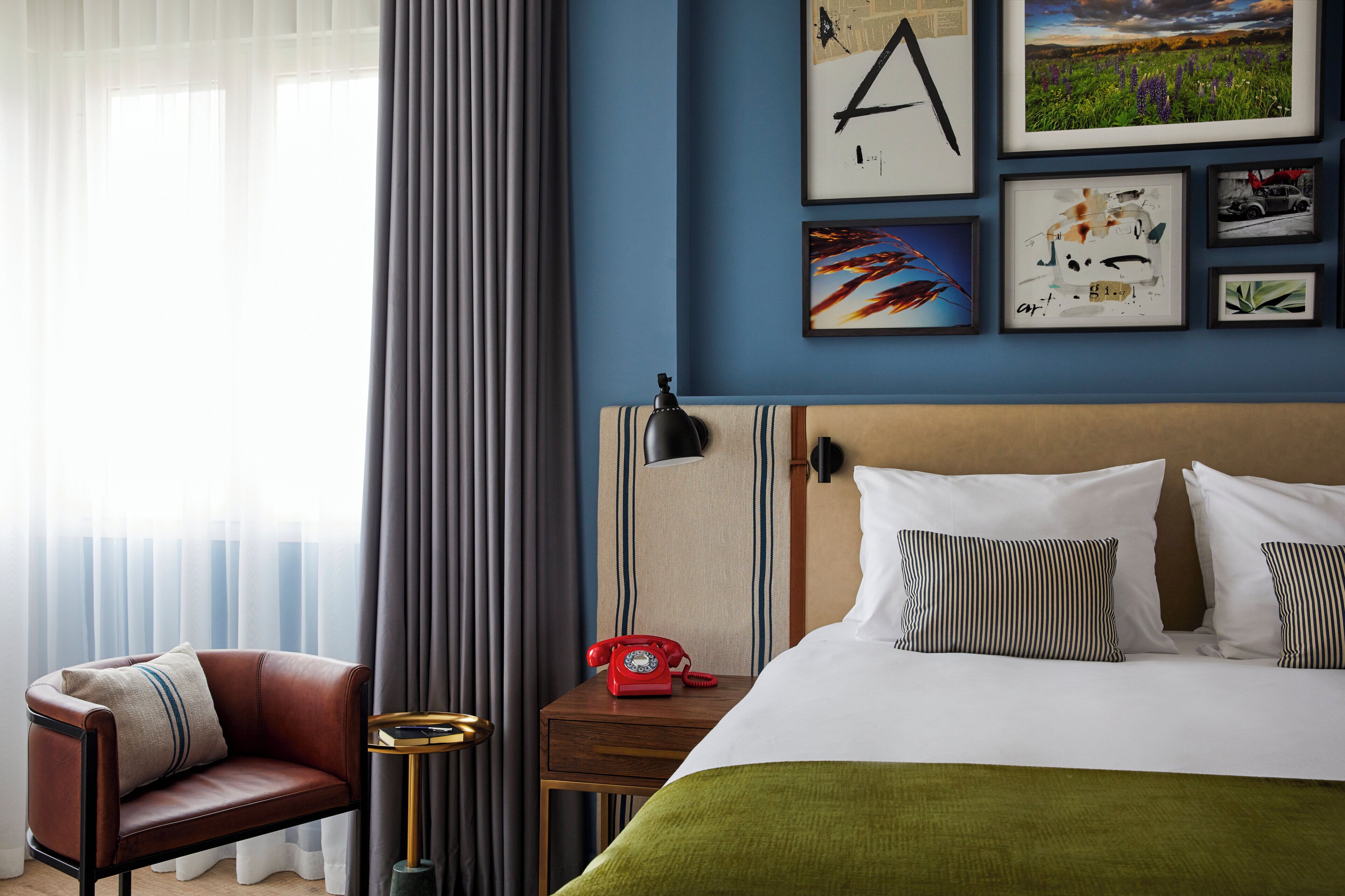 Una habitación de hotel con una cama, una silla, una mesita de noche con un teléfono rojo y una pared con cuadros.