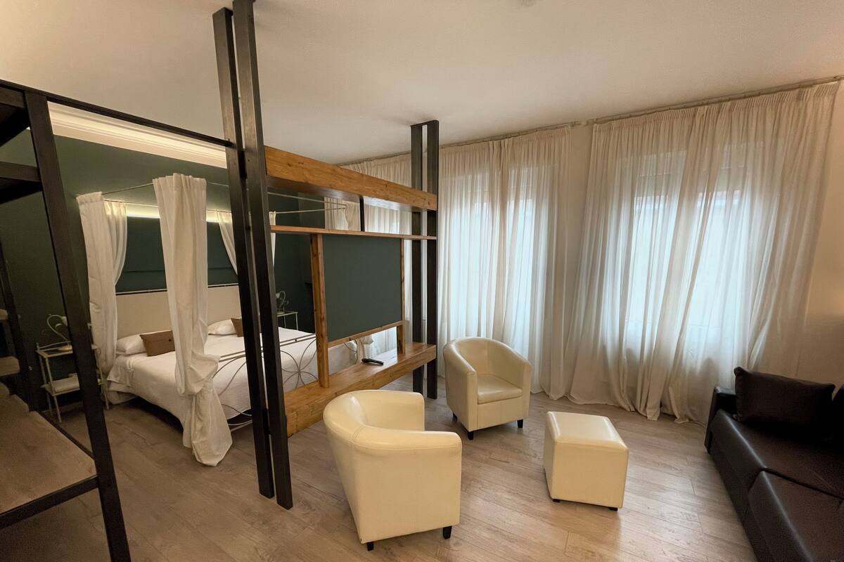 Ein modernes Hotelzimmer mit einem Bett, einer Sitzecke mit Sessel und Hocker, einer Holzbank und einem großen Fenster mit Vorhängen.