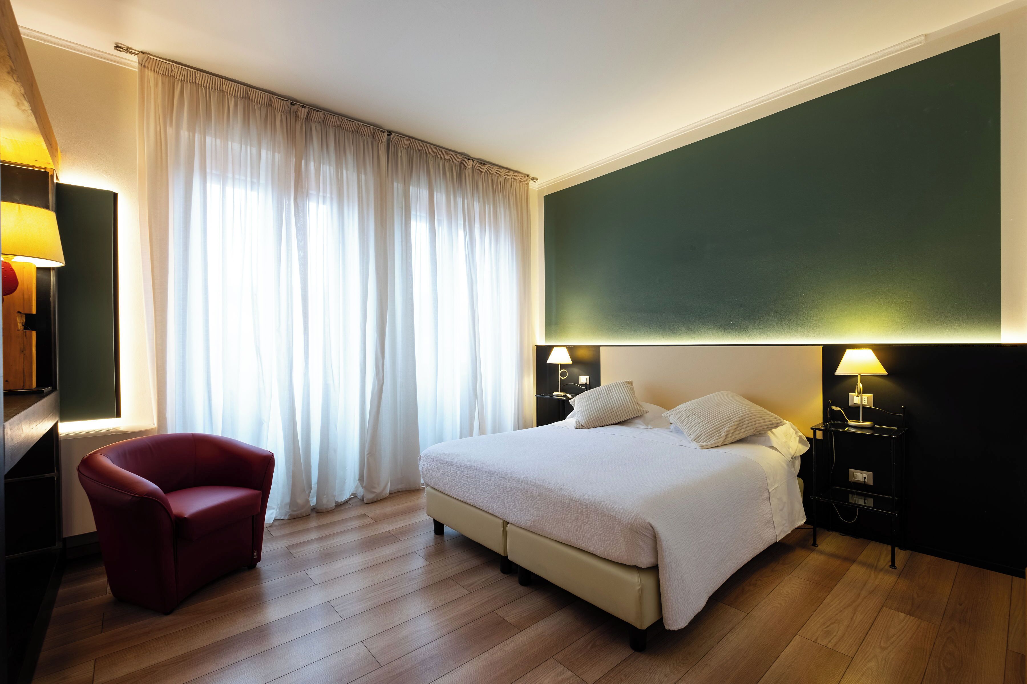 Ein Hotelzimmer mit einem Bett, einem roten Sessel, einem Nachttisch mit Lampe und einem großen Fenster mit Vorhängen.