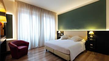 Ein Hotelzimmer mit einem Bett, einem roten Sessel, einem Nachttisch mit Lampe und einem großen Fenster mit Vorhängen.