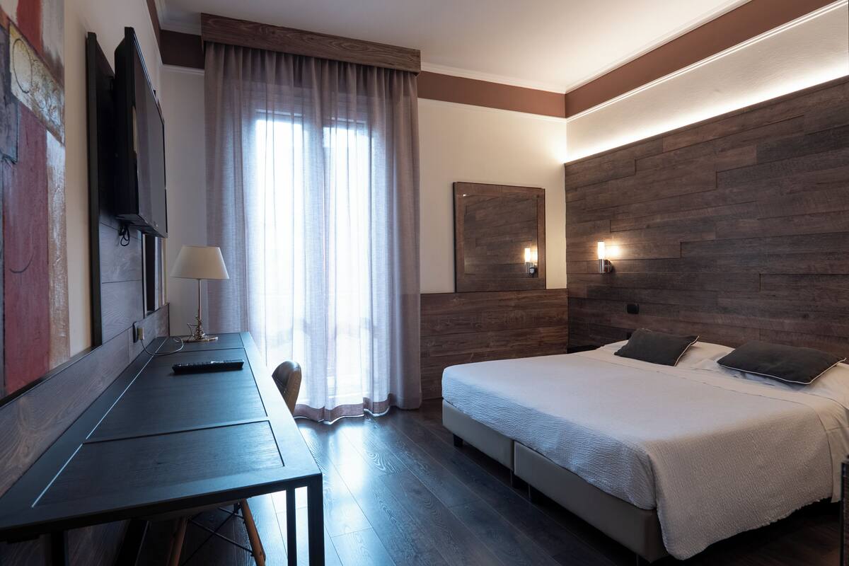 Ein Hotelzimmer mit Bett, Schreibtisch, Sessel, Fernseher und einem Fenster mit Vorhängen.