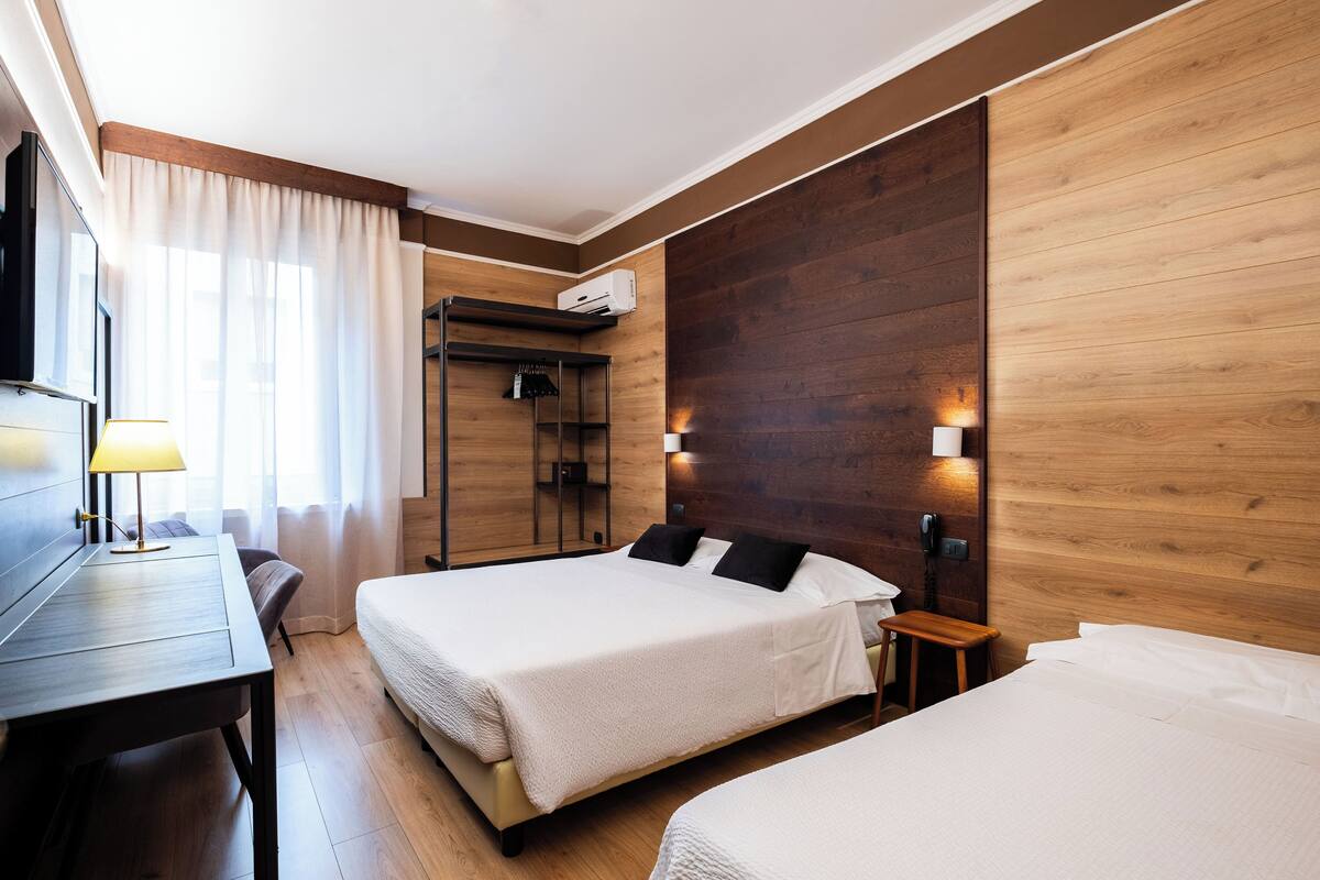 Ein Hotelzimmer mit zwei Betten, einem Schreibtisch, einem Sessel, einem Fernseher, einer Lampe und einer Holzverkleidung an der Wand.