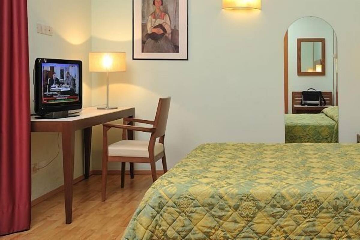 A hotel room with a bed, a desk with a television, a chair, a bedside table with books, and a mirror.