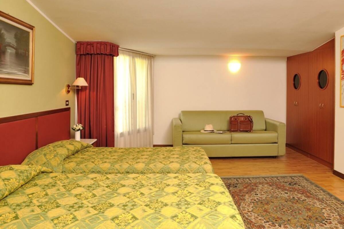 A hotel room with a bed, a green sofa, a wooden wardrobe, and a painting on the wall.