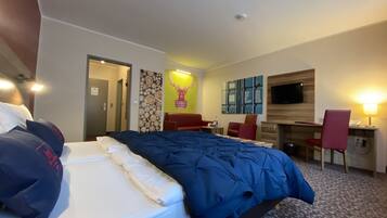 A hotel room with a bed, a desk, a chair, a TV, and a wall with artwork.