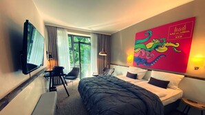 Una habitación de hotel con una cama grande, un escritorio con una silla, un televisor montado en la pared y una obra de arte colorida en la pared.