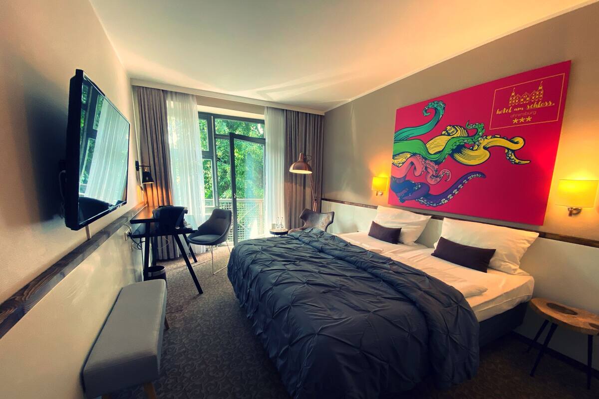 Una habitación de hotel con una cama grande, un escritorio con una silla, un televisor montado en la pared y una obra de arte colorida en la pared.