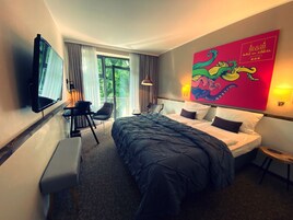 Ein Hotelzimmer mit einem großen Bett, einem Schreibtisch mit Stuhl, einem an der Wand montierten Fernseher und einem farbenfrohen Kunstwerk.