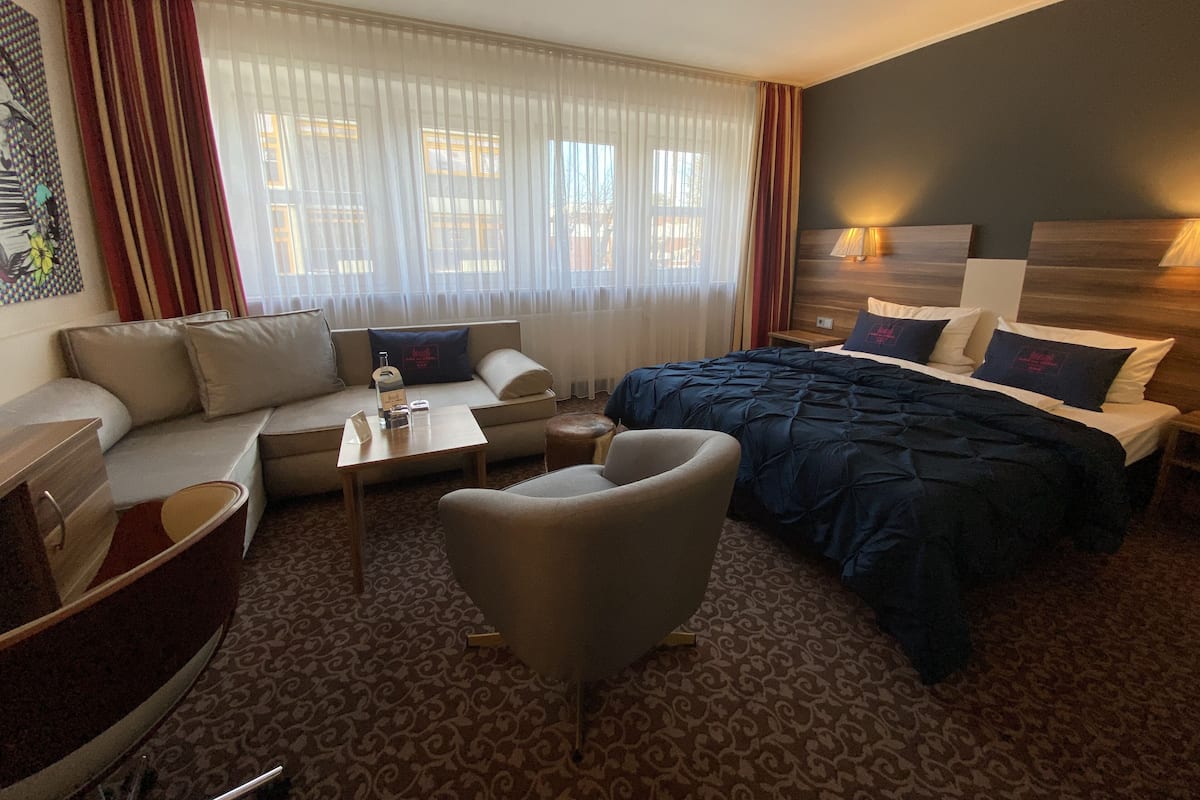 Habitación de hotel con cama, sofá, sillón, mesita y ventana con cortinas.