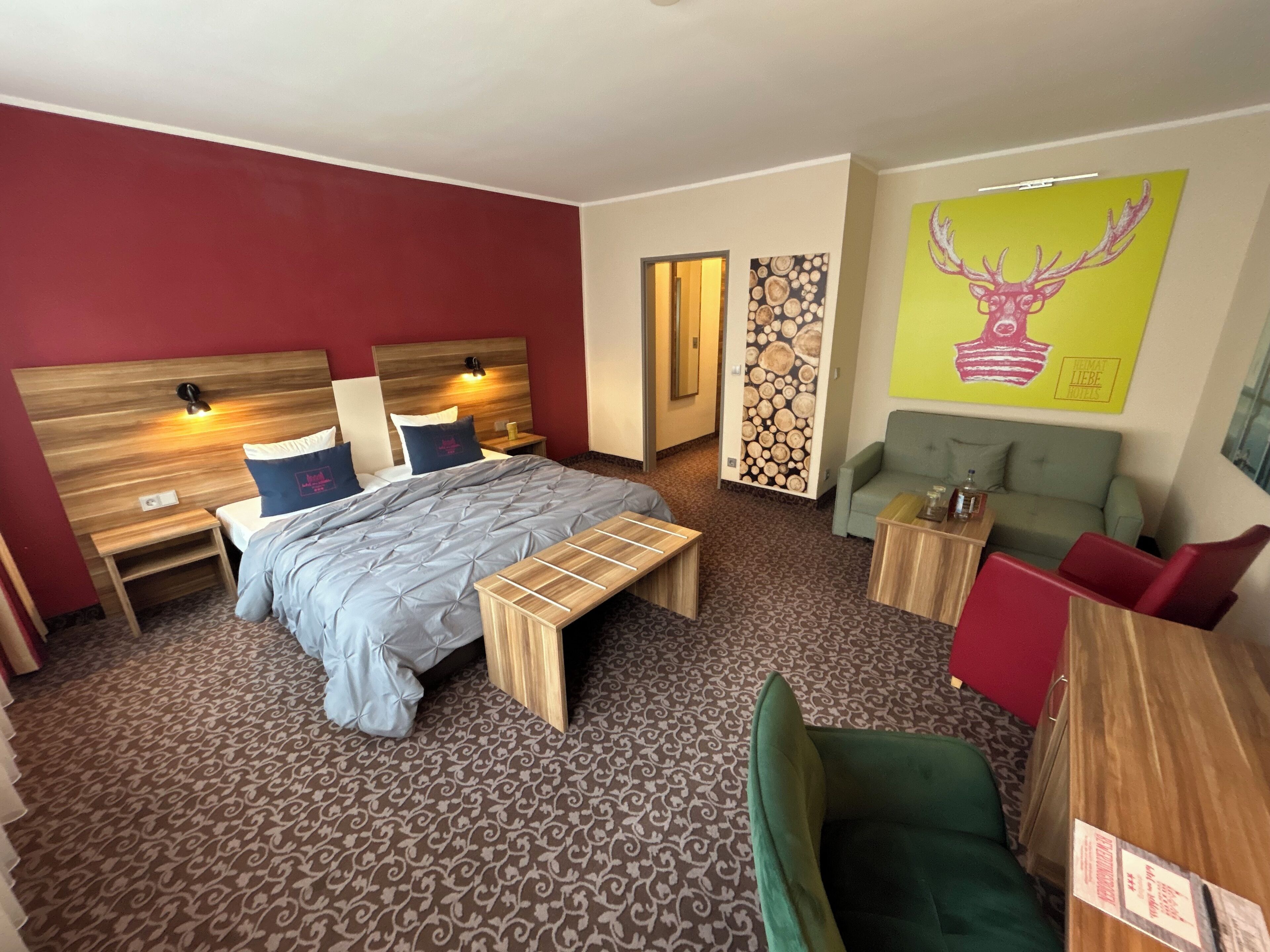 Une chambre d’hôtel comprenant un lit, des tables de chevet, un banc, un canapé, une petite table et un mur orné d’un tableau représentant un cerf.