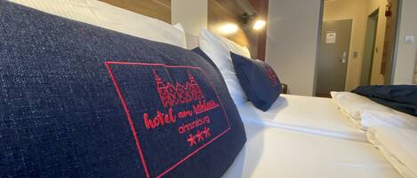 A hotel bed with a dark blue pillow featuring red embroidery of a hotel name and a city skyline.