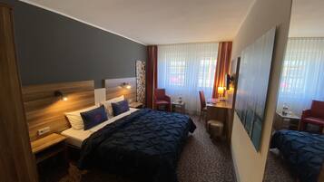 Habitación de hotel con una cama grande, un escritorio con una silla, una mesita de noche con lámpara y un espejo.