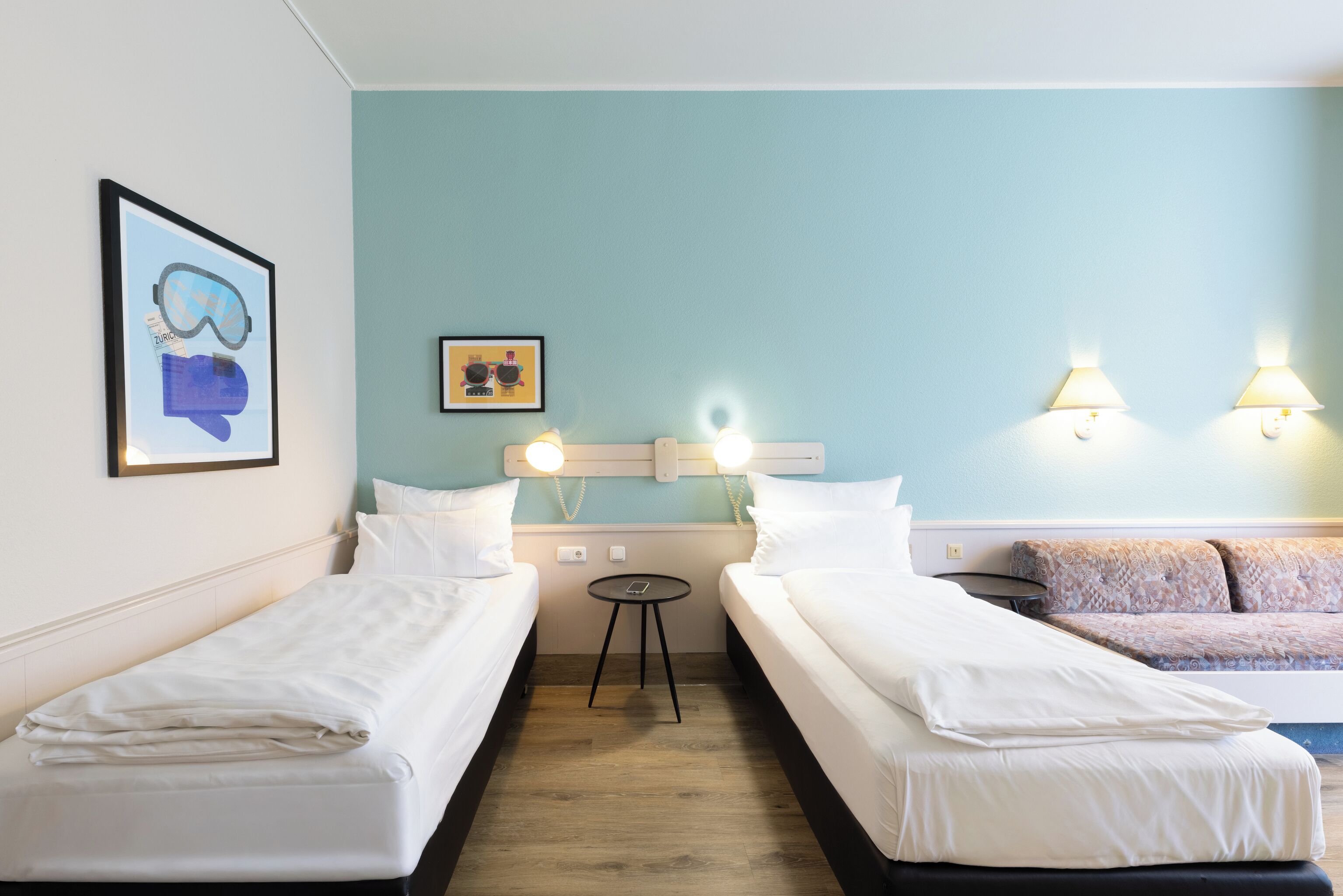 A hotel room with two single beds, a small table, a sofa, and wall-mounted lights.