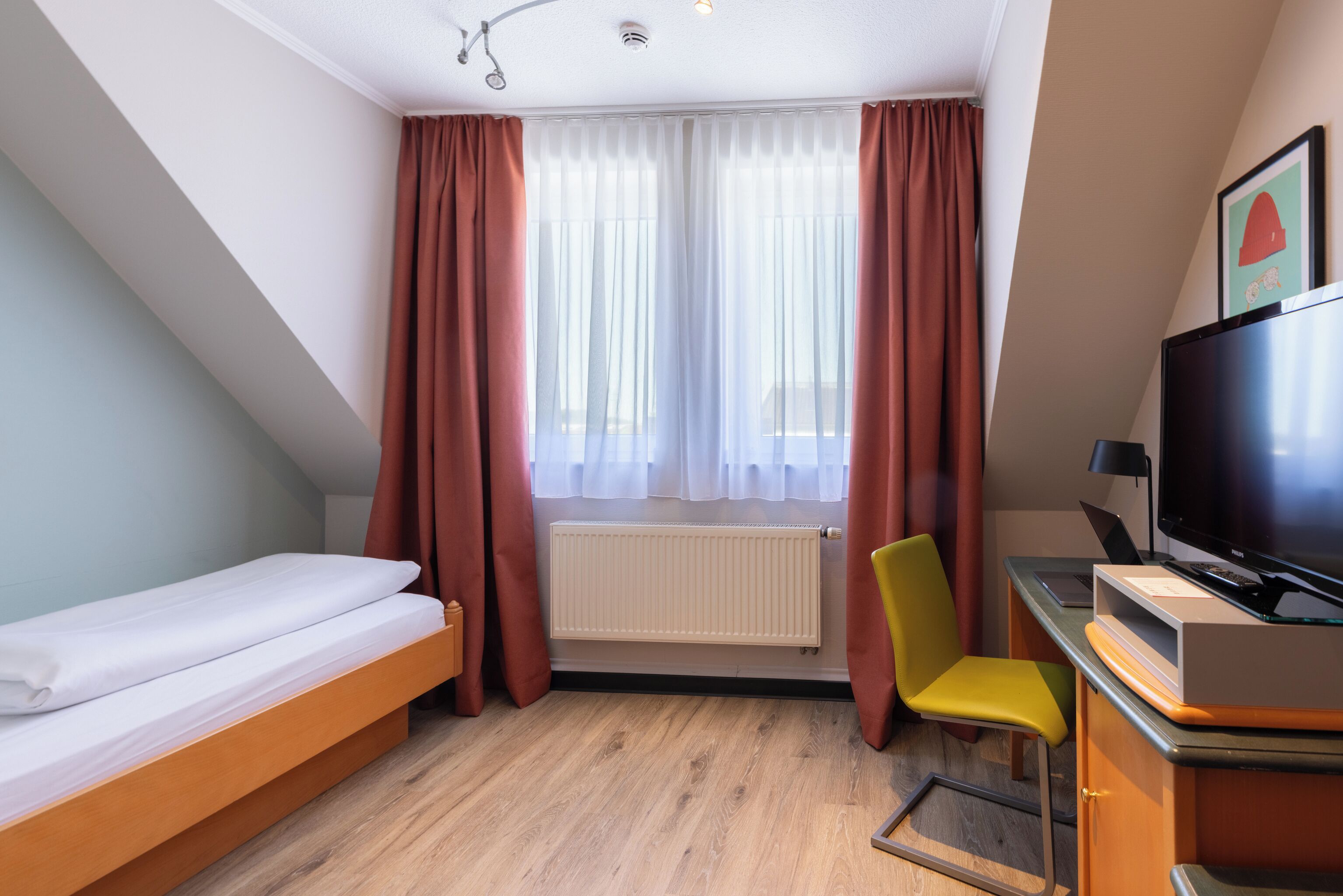 Habitación de hotel con cama, escritorio con computadora, silla, televisor y ventana con cortinas.