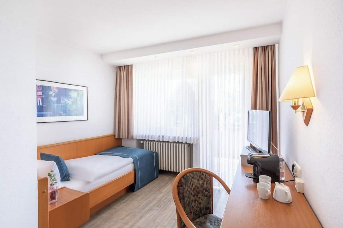 Une chambre d’hôtel avec un lit, un bureau avec une lampe, une chaise, une télévision et une fenêtre avec des rideaux.