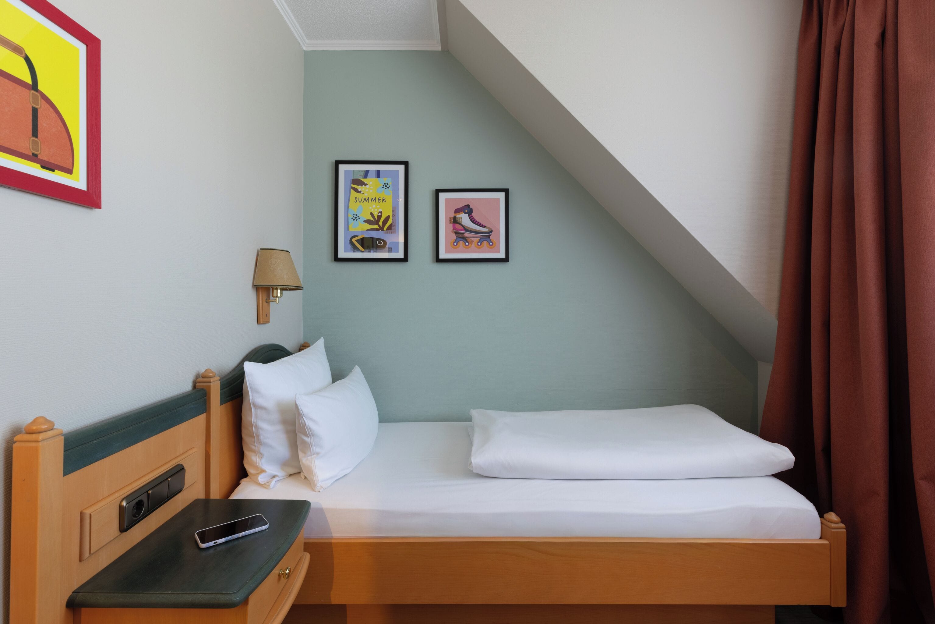 Een eenpersoonsbed met witte lakens, een nachtkastje met een lamp, en twee ingelijste foto's aan de muur.