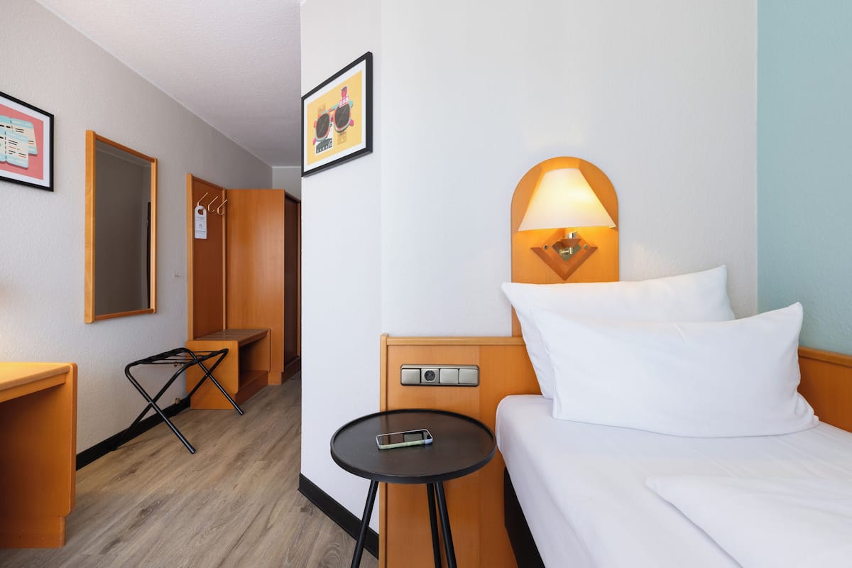 Une chambre d’hôtel comprenant un lit, une table de chevet, une petite table ronde, une lampe fixée au mur, un porte-manteau et un tableau encadré accroché au mur.
