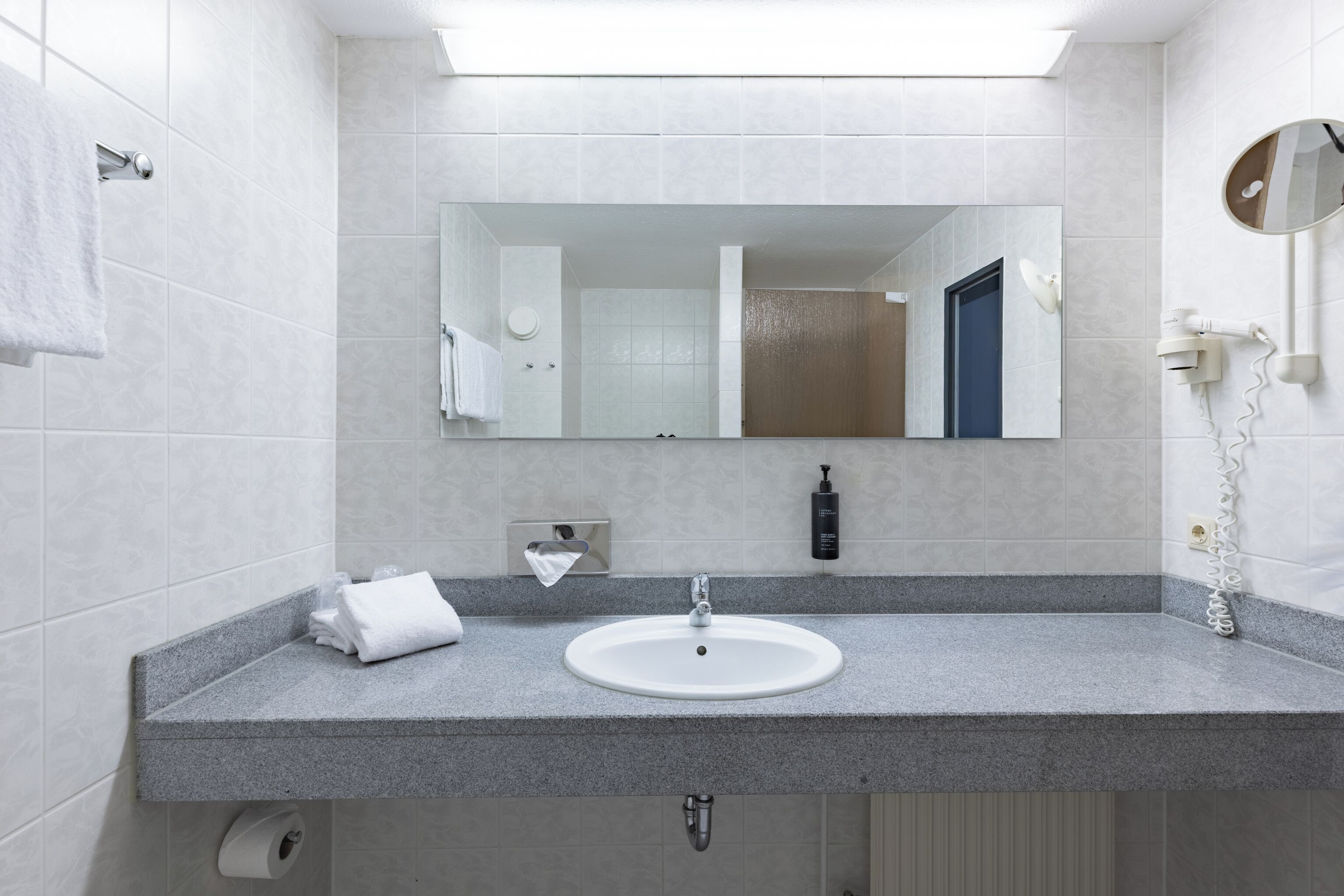 Un baño con lavamanos blanco, un espejo, toallero y secador de pelo.