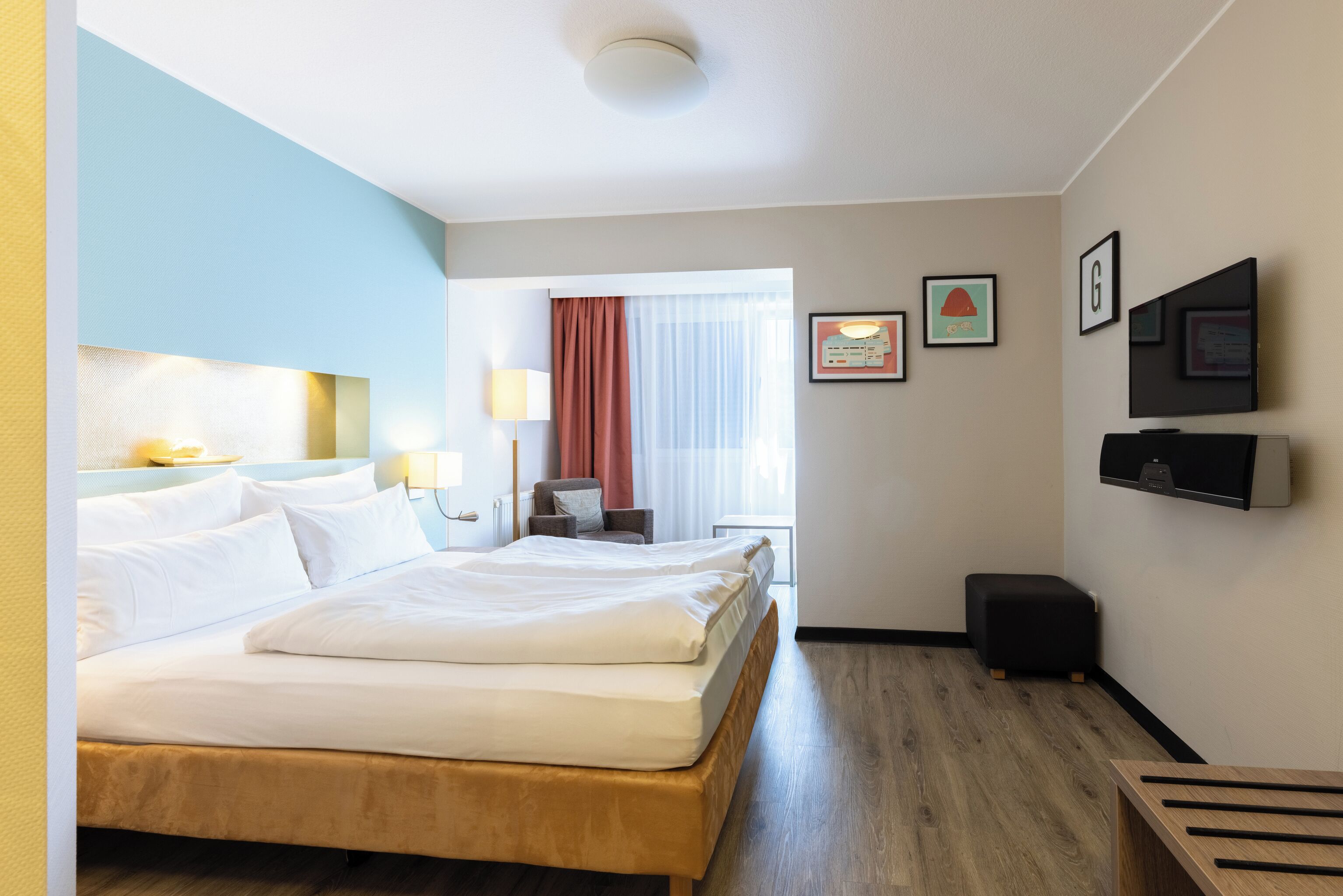 Une chambre d’hôtel comprenant un lit, des tables de chevet, un bureau, une chaise, une fenêtre avec des rideaux et des œuvres d’art accrochées au mur.