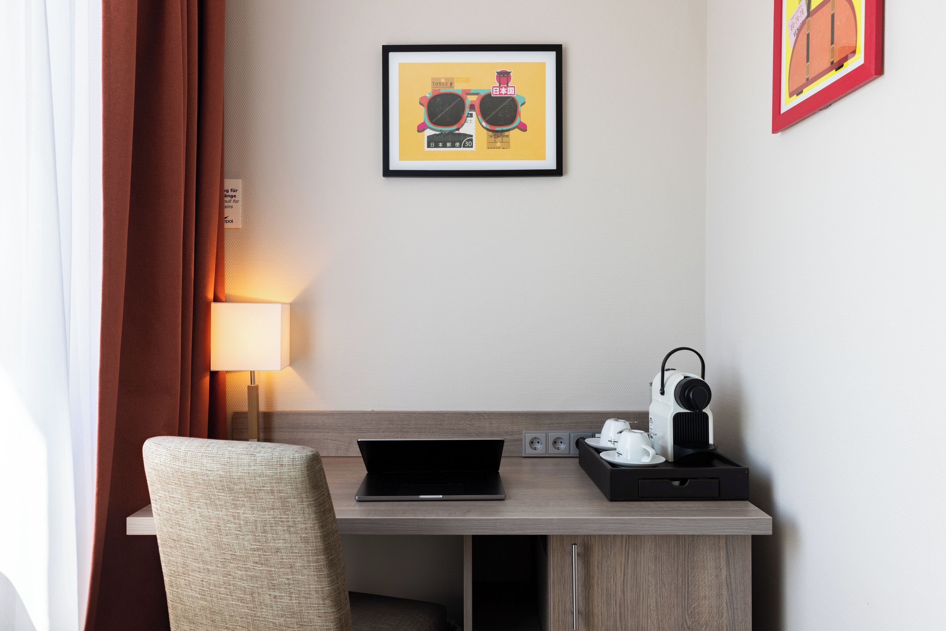 A hotel room with a desk, laptop, lamp, and a framed picture on the wall.