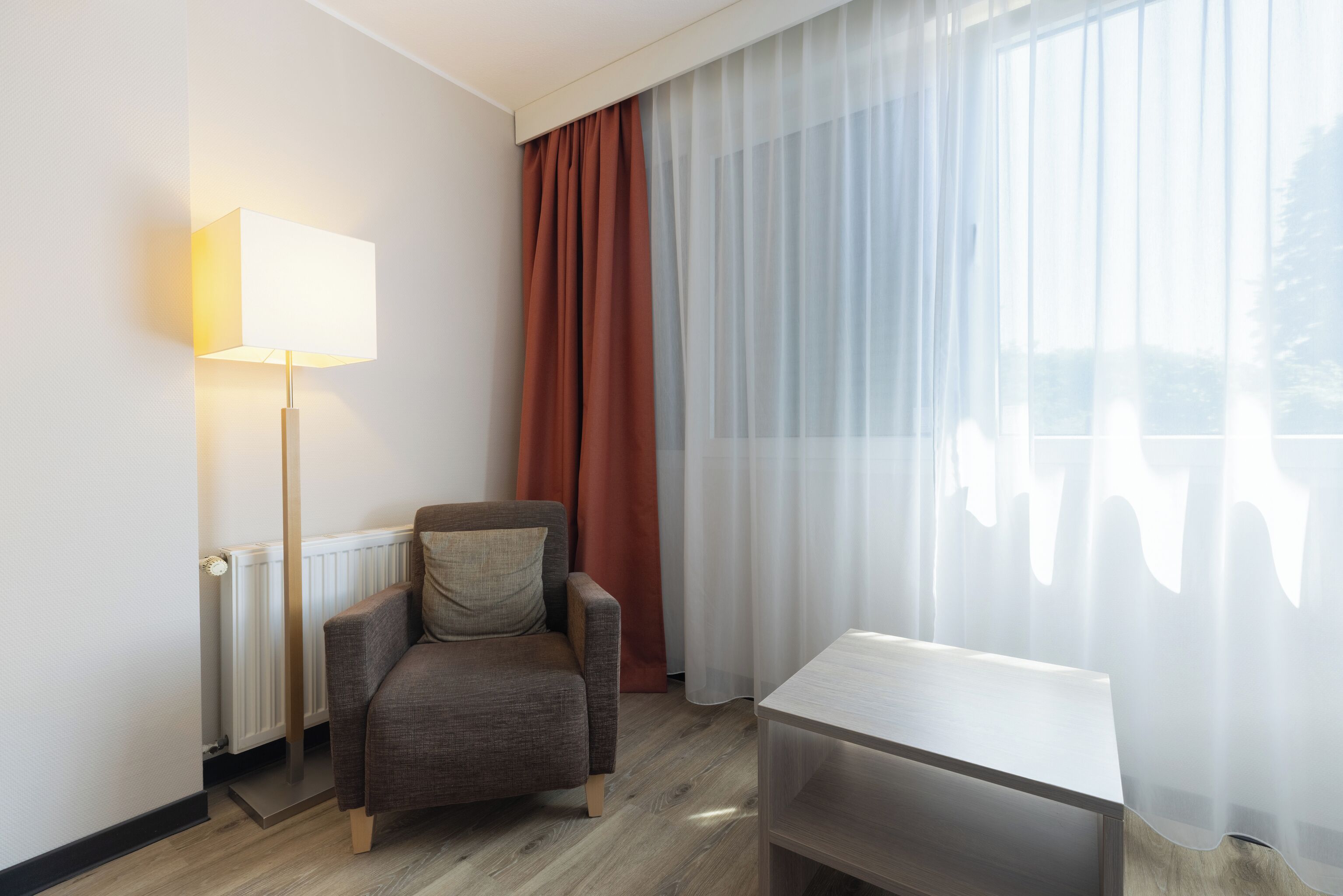 Habitación de hotel con una silla, una mesa, una lámpara y una ventana con cortinas traslúcidas.