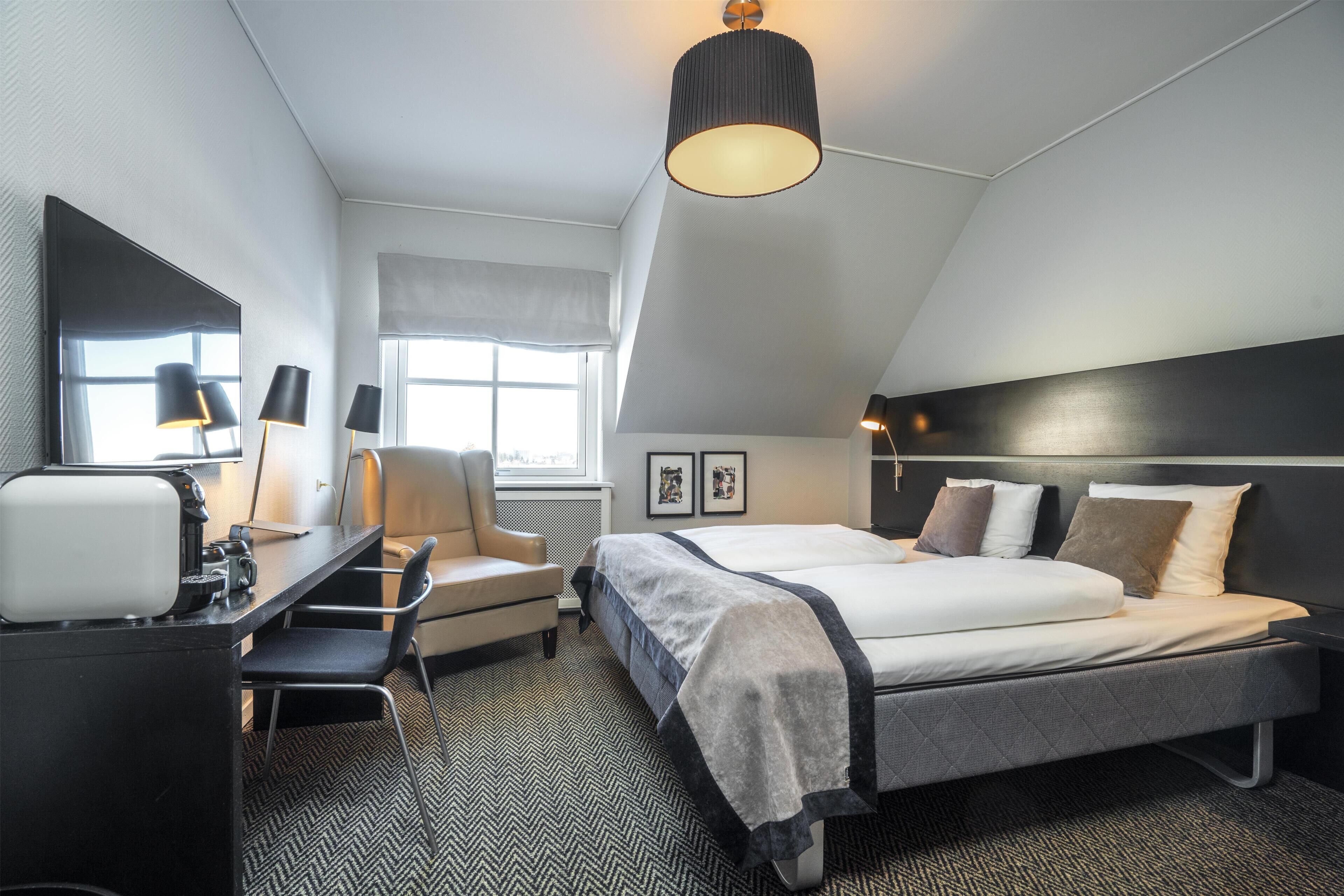 A hotel room with a bed, desk, chair, and a large window.