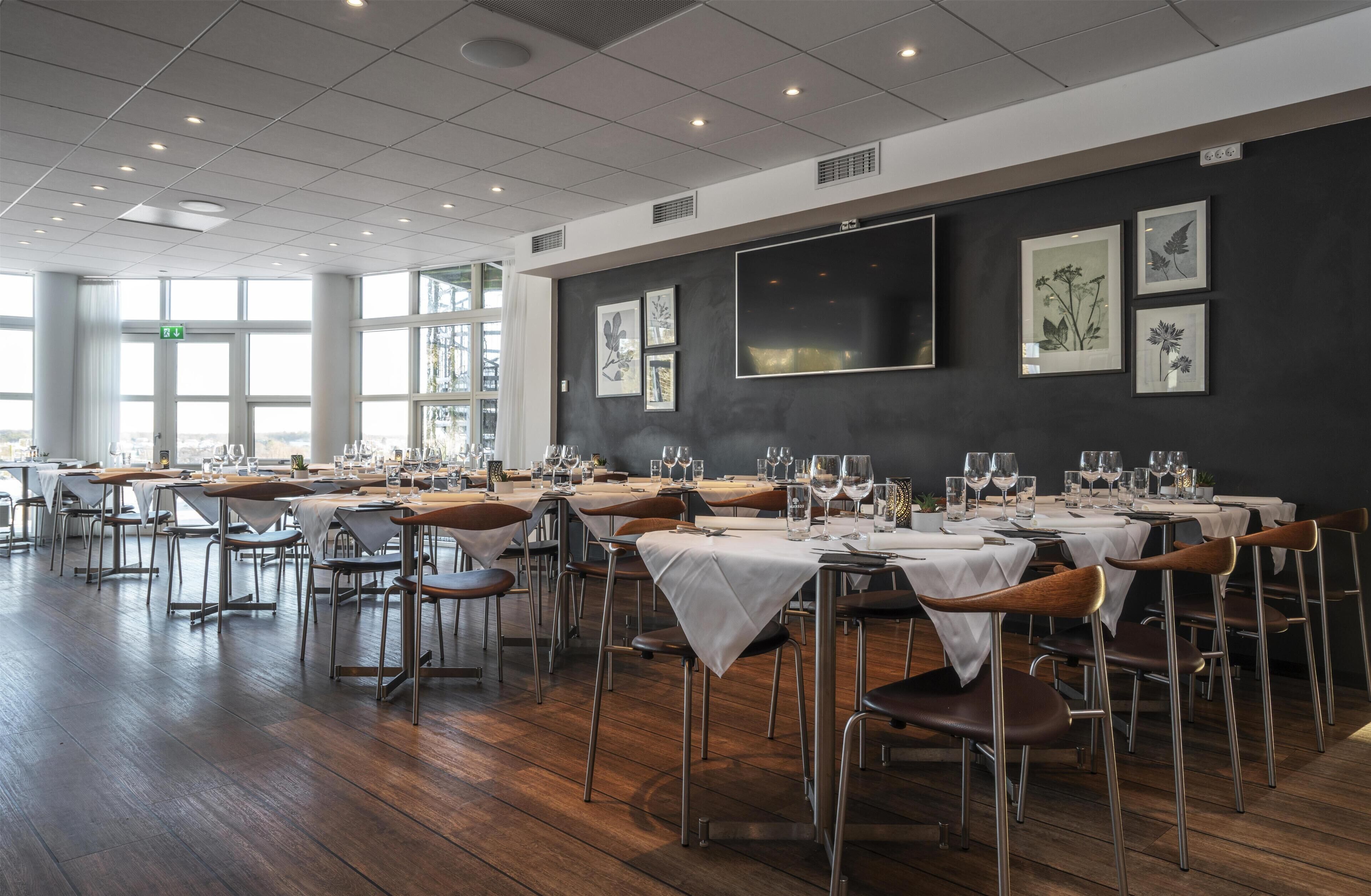 Un restaurante con varias mesas dispuestas para comer, con un interior moderno, piso de madera y una pared oscura decorada con impresiones botánicas enmarcadas.