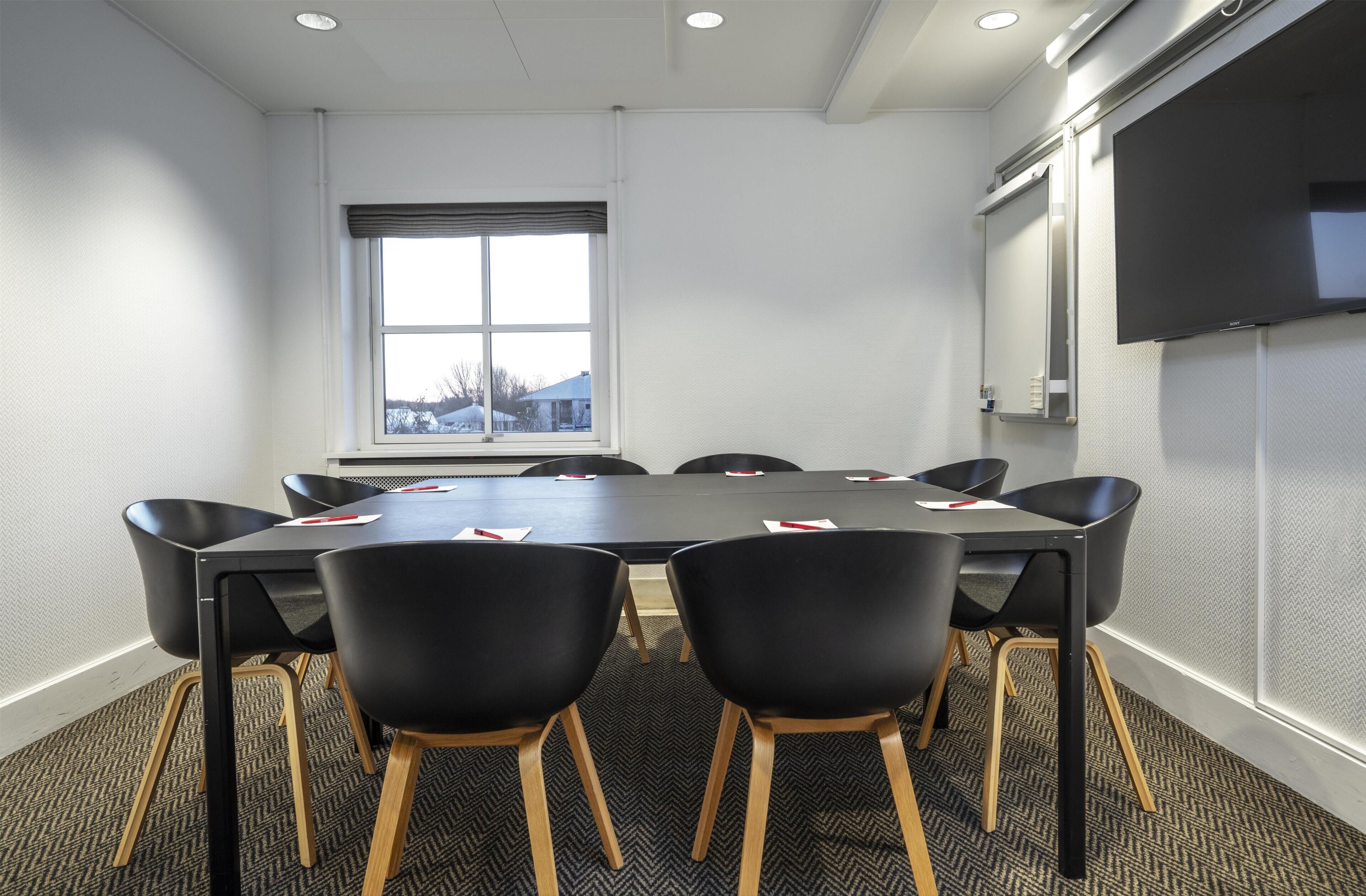 A conference room with a large table, six chairs, a TV screen, and a window with a view of buildings.