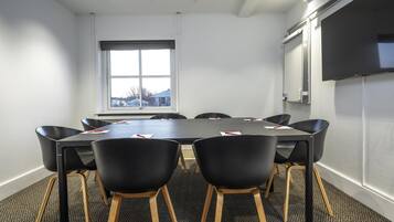 A conference room with a large table, six chairs, a TV screen, and a window with a view of buildings.