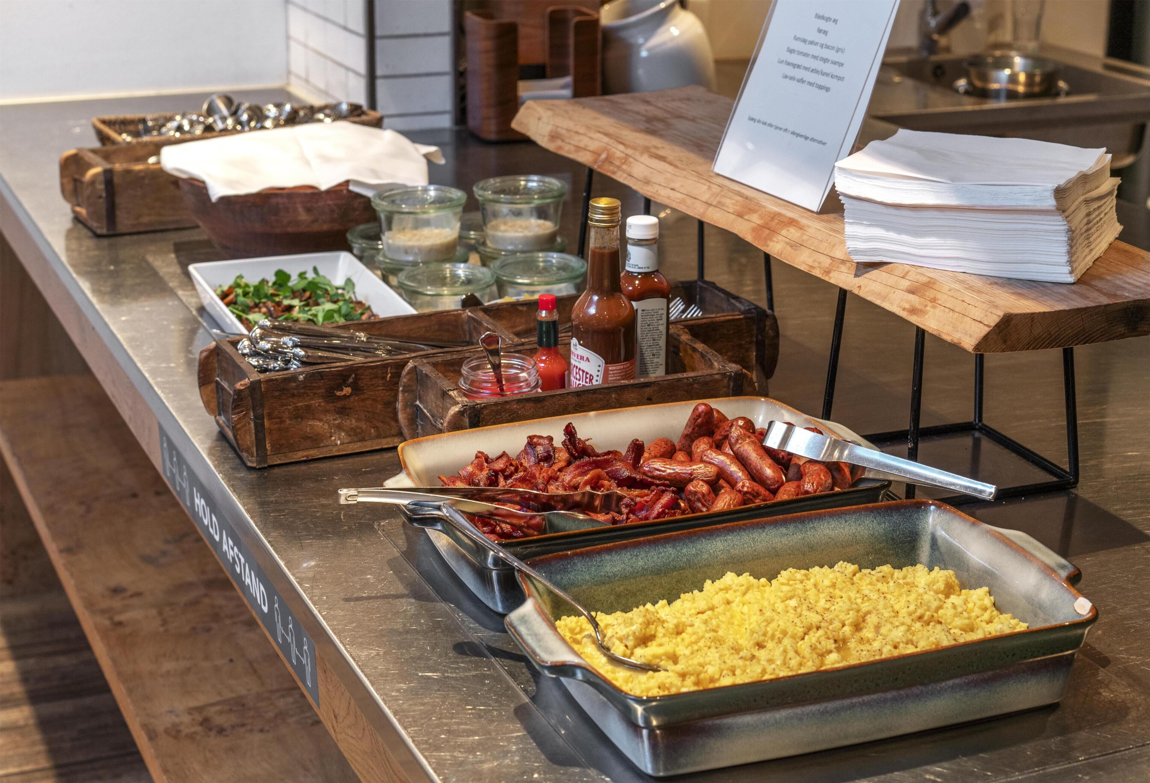 Una mesa de buffet con diferentes alimentos, incluyendo tocino, salchichas y un plato gratinado.