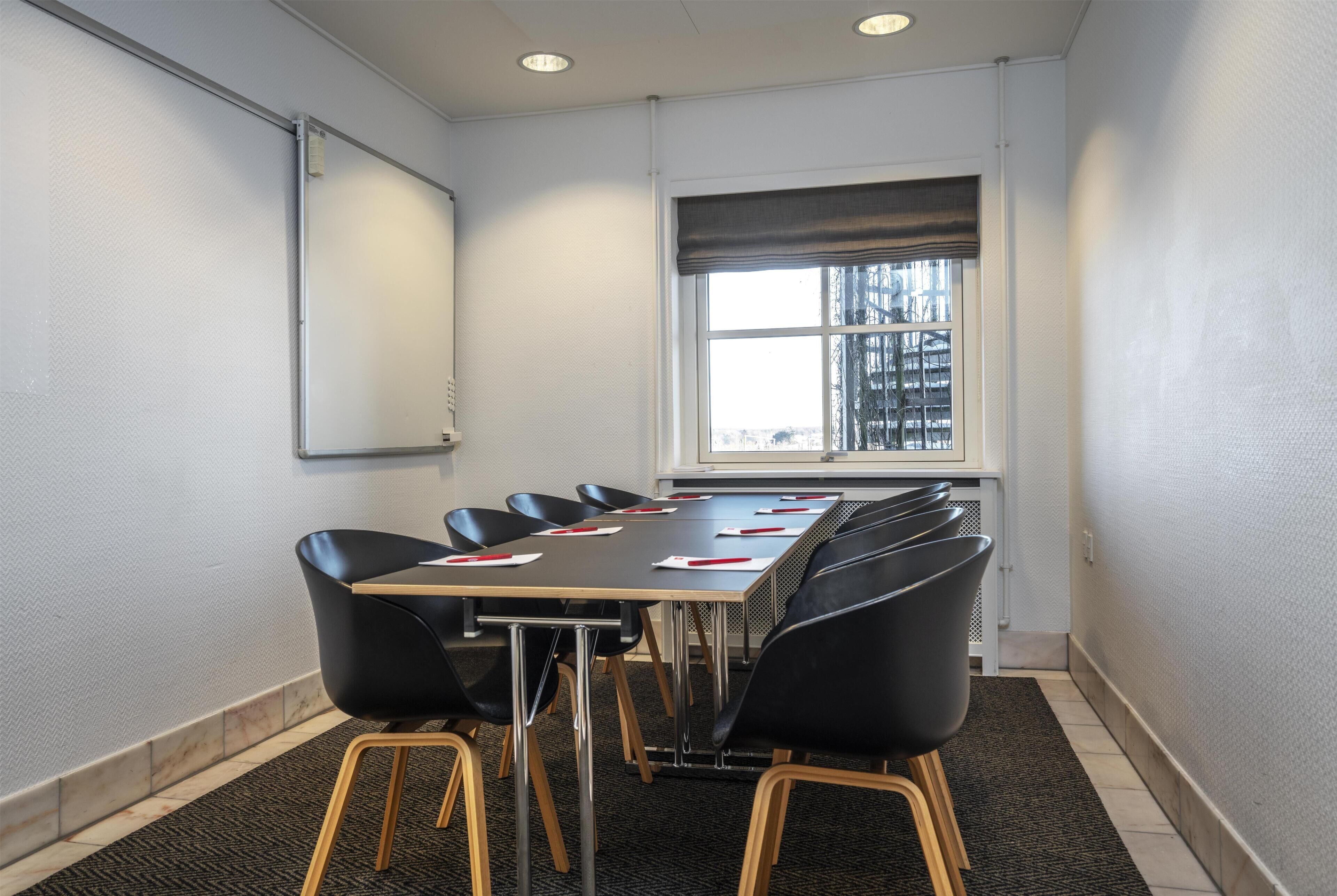 Une salle de conférence avec une longue table, des chaises noires, un tableau blanc et une fenêtre donnant sur des immeubles.