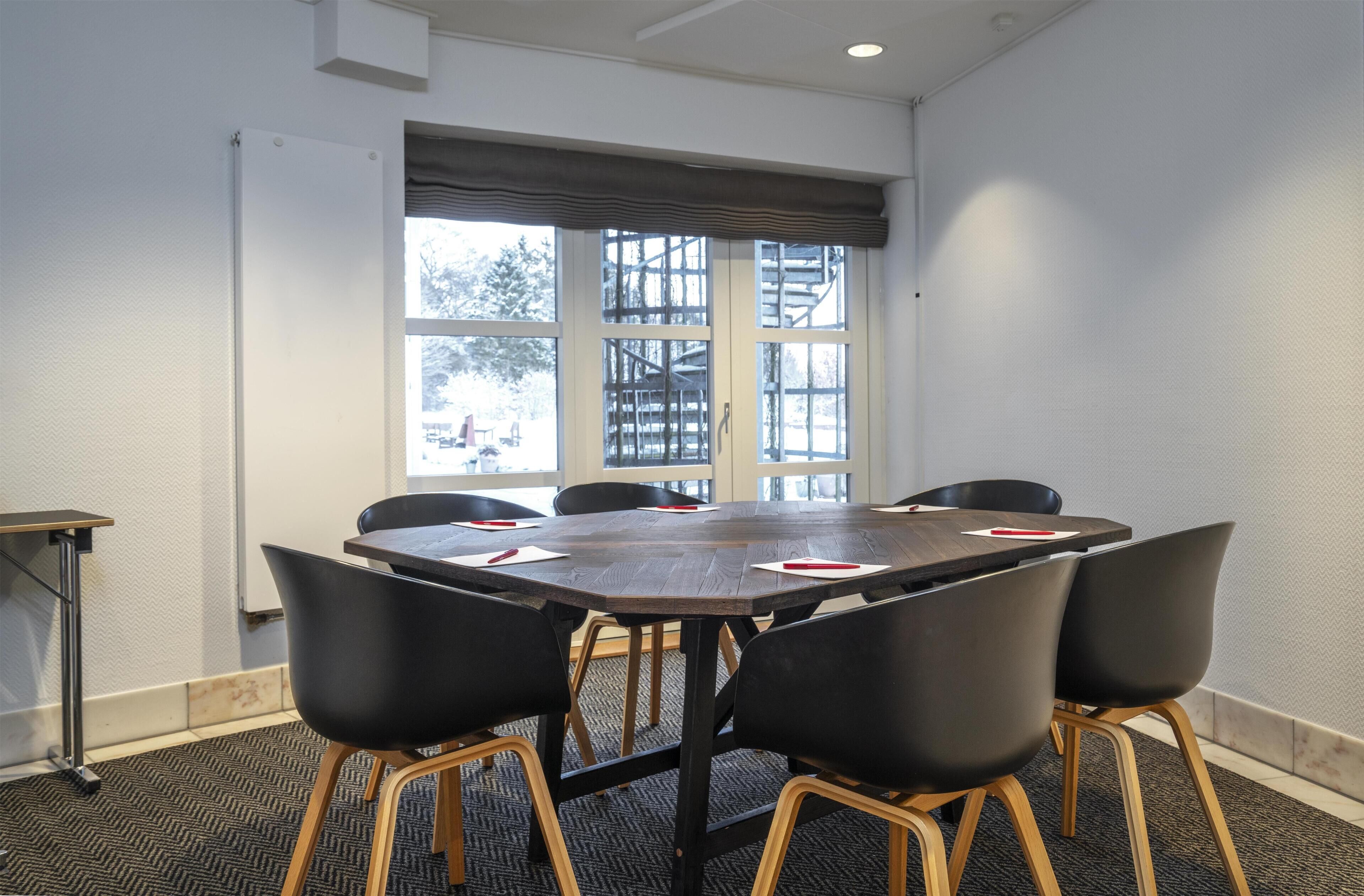 Een vergaderruimte met een grote tafel, zes stoelen en een raam met uitzicht op bomen en een gebouw.