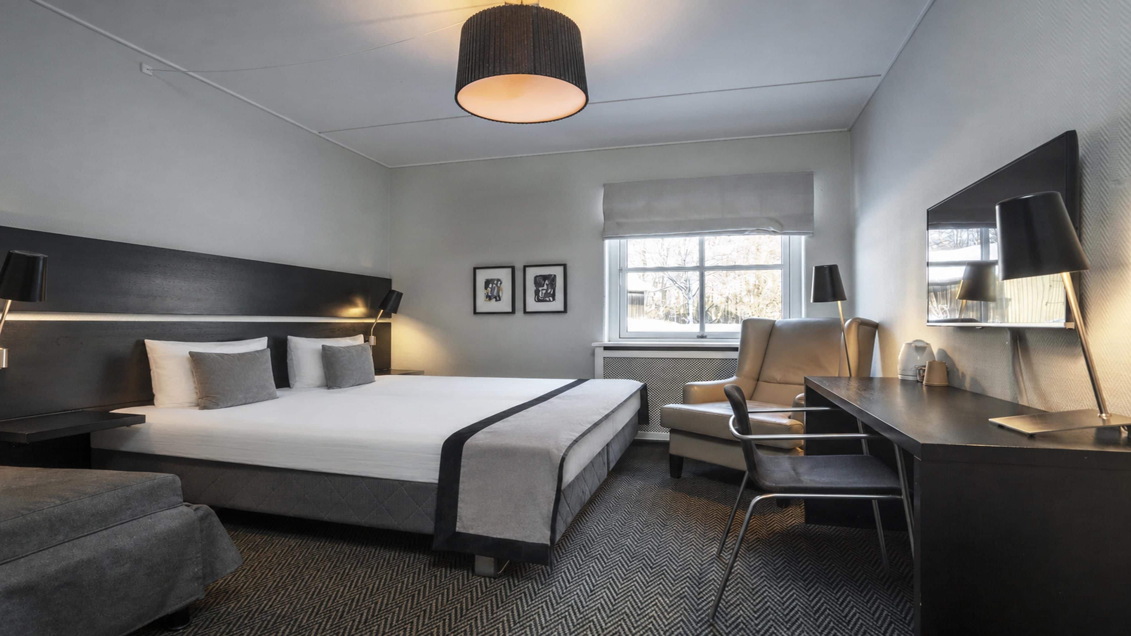 A hotel room with a bed, desk, chair, and a large window.