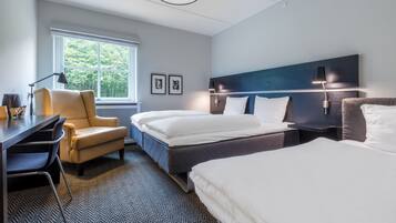 A hotel room with a desk, two beds, a chair, a window with a view of trees, and framed pictures on the wall.
