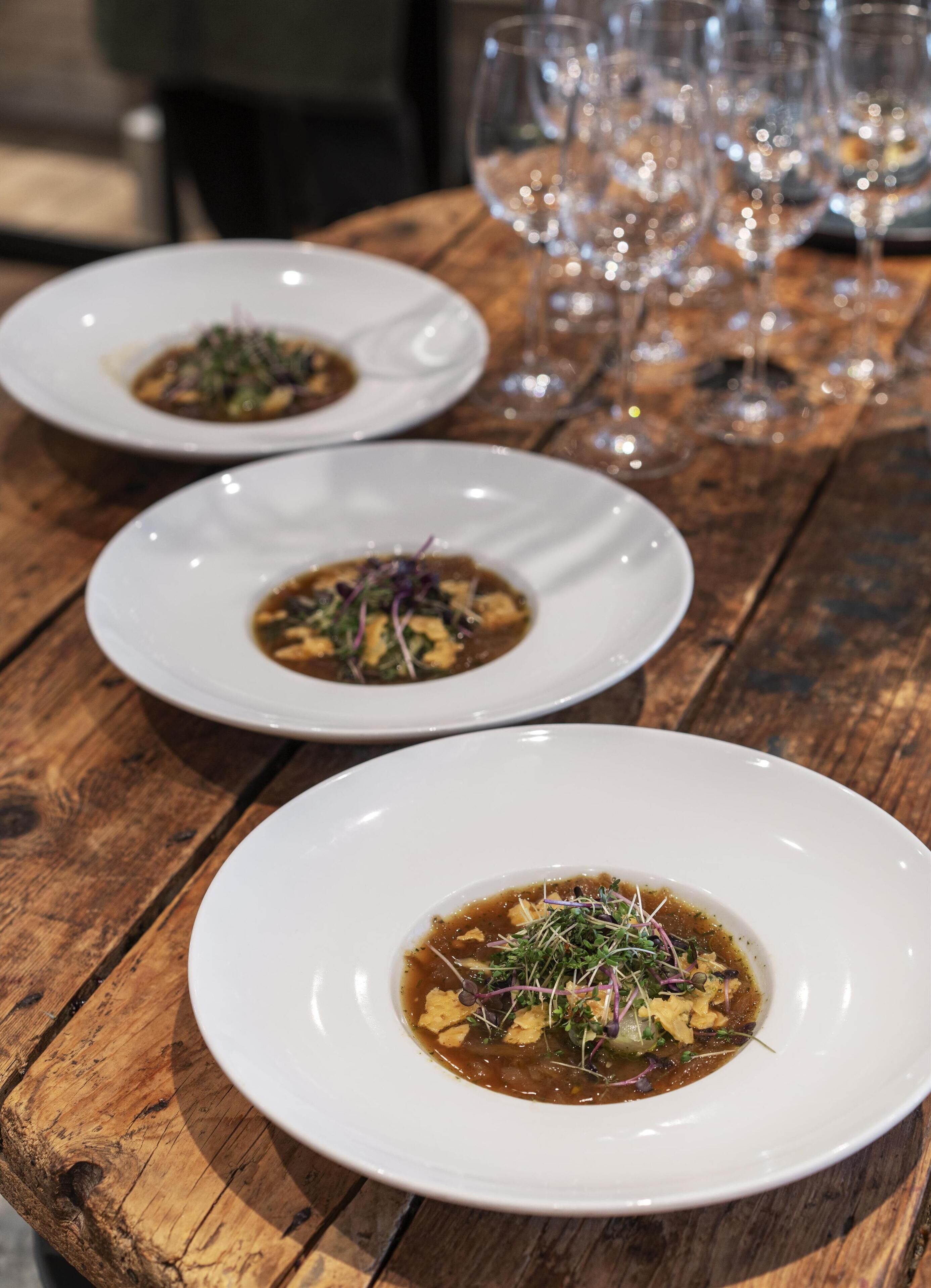 Trois assiettes blanches remplies de nourriture sont posées sur une table en bois, avec des verres à vin en arrière-plan.