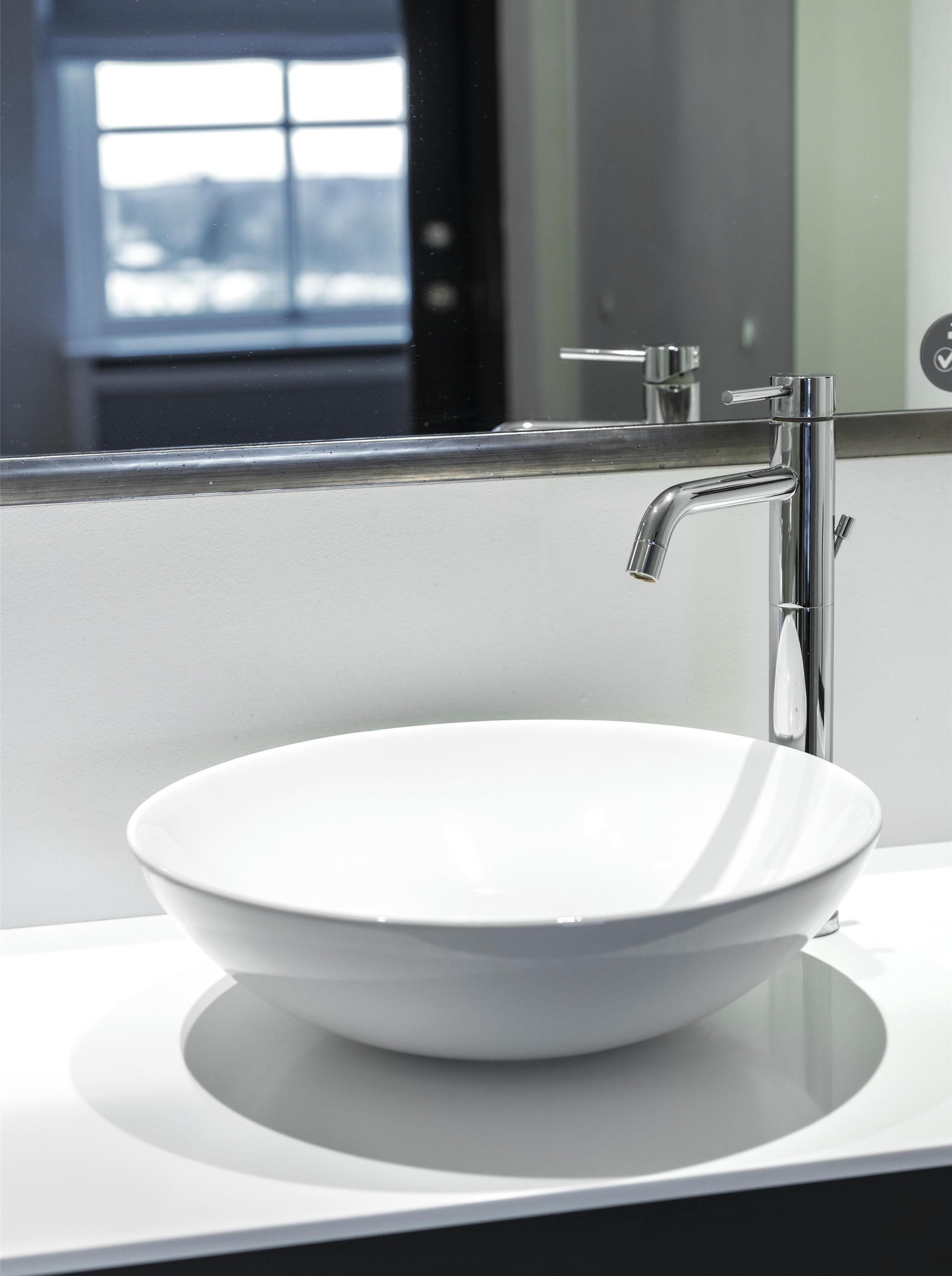 A modern bathroom with a white sink, chrome faucet, and a mirror reflecting a window.