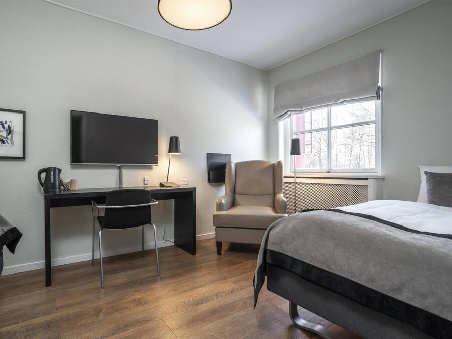 A hotel room with a bed, a desk with a chair, a television, and a window with a view of trees.