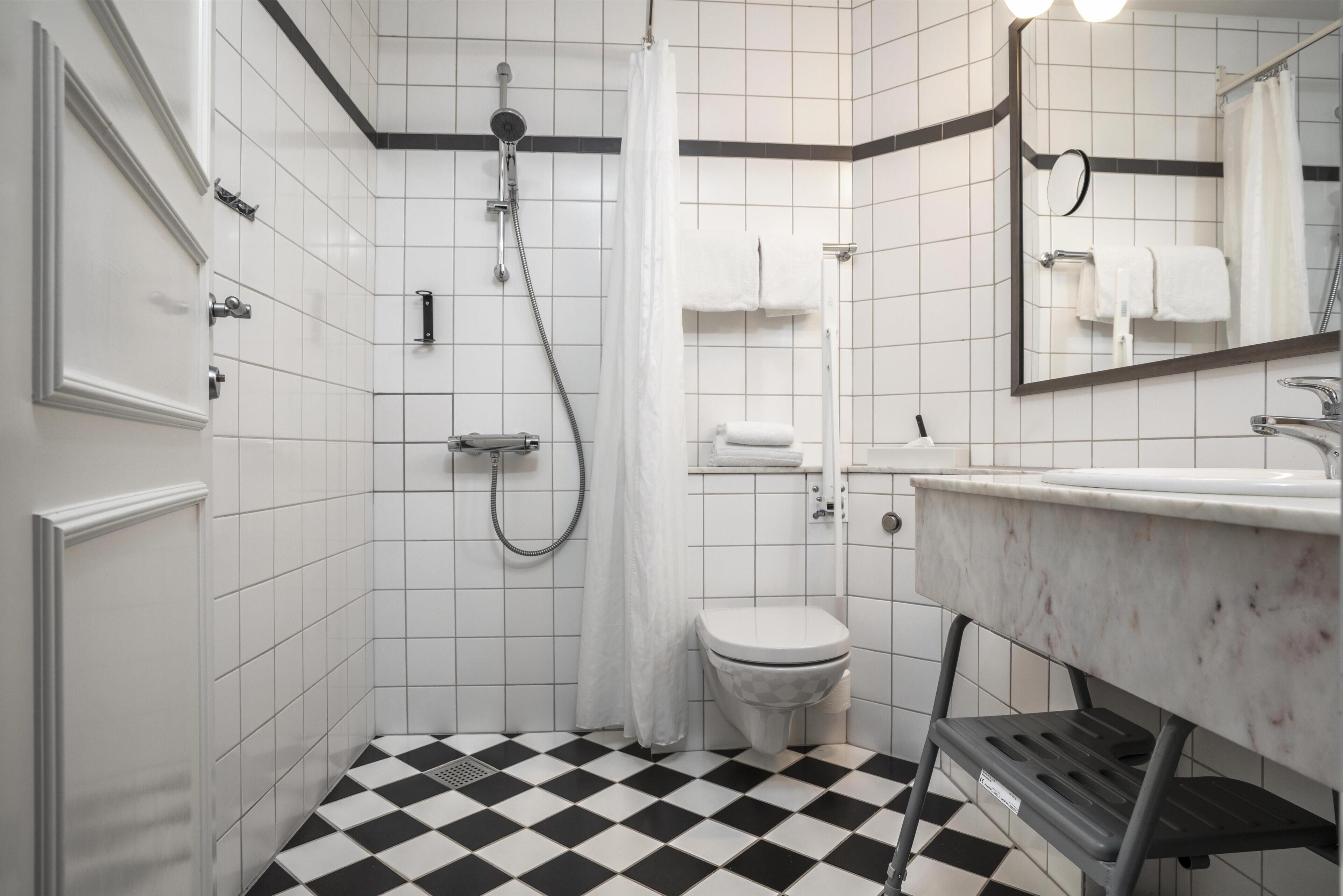 Un baño con piso a cuadros blanco y negro, inodoro blanco, ducha con cortina y lavamanos con espejo arriba.