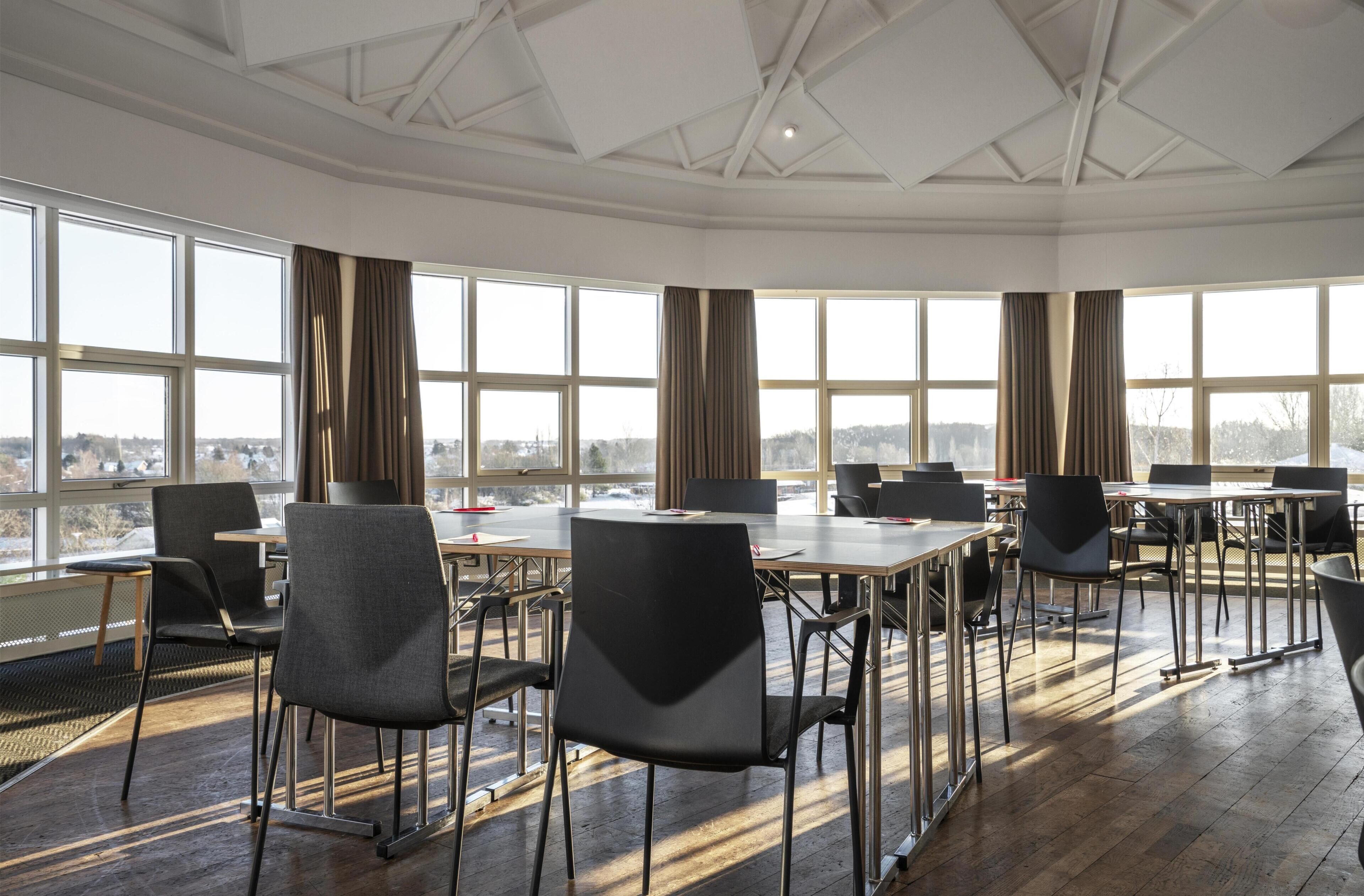 Un salón de conferencias con varias mesas y sillas, ventanas amplias y vista panorámica de la ciudad.