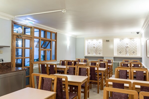 A dining area with wooden tables and chairs, a kitchen area with a view through a glass door, and decorative wall sconces. - Nayland Hotel (London)