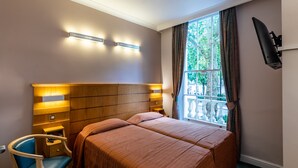 A hotel room with a wooden headboard, two beds, a chair, a TV mounted on the wall, and a window with curtains. - Nayland Hotel (London)