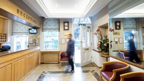A reception area with a wooden reception desk, a clock, and a floral arrangement. - Nayland Hotel (London)