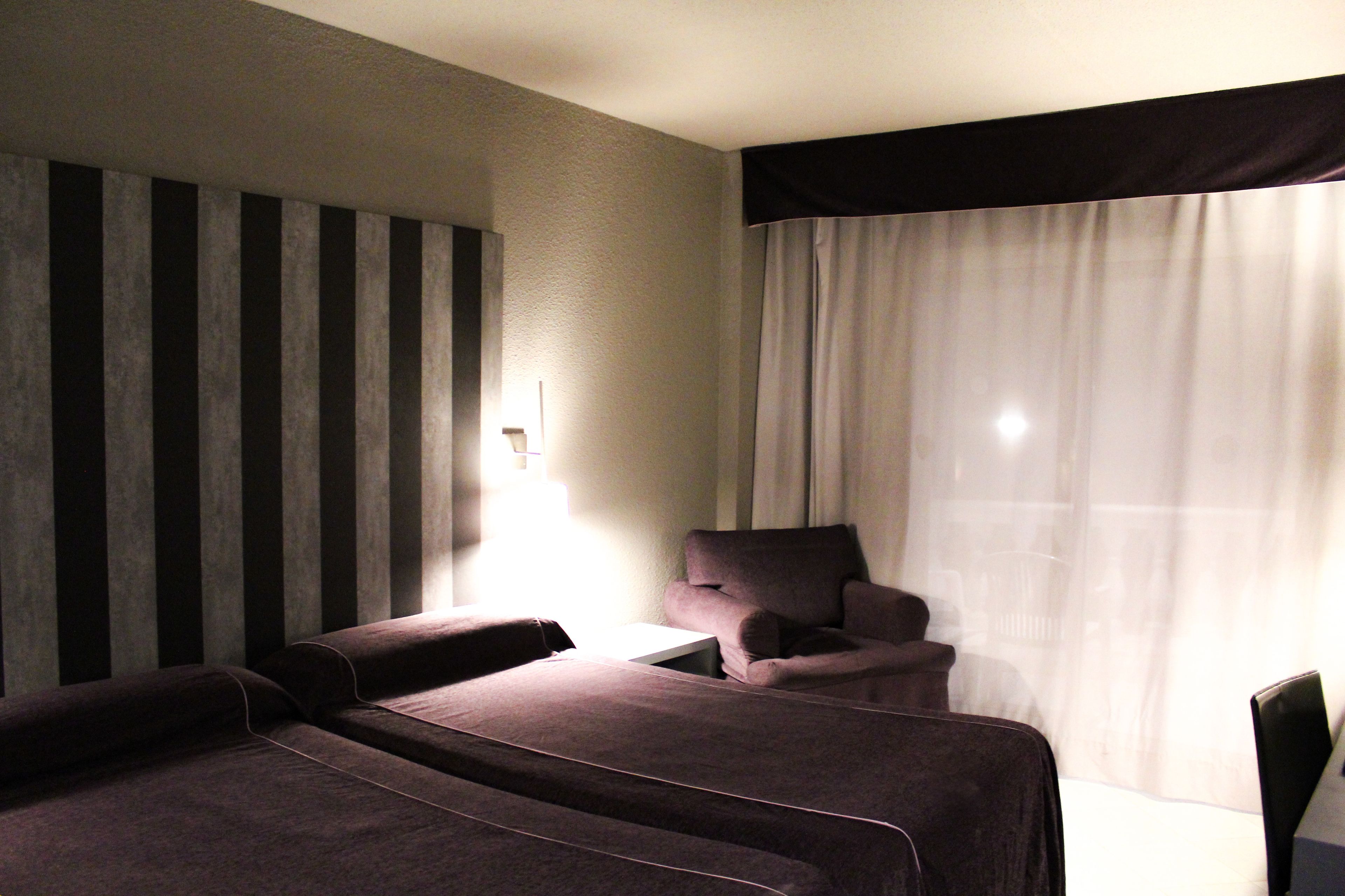 Habitación de hotel con dos camas, una silla, una mesita y una ventana con cortinas.