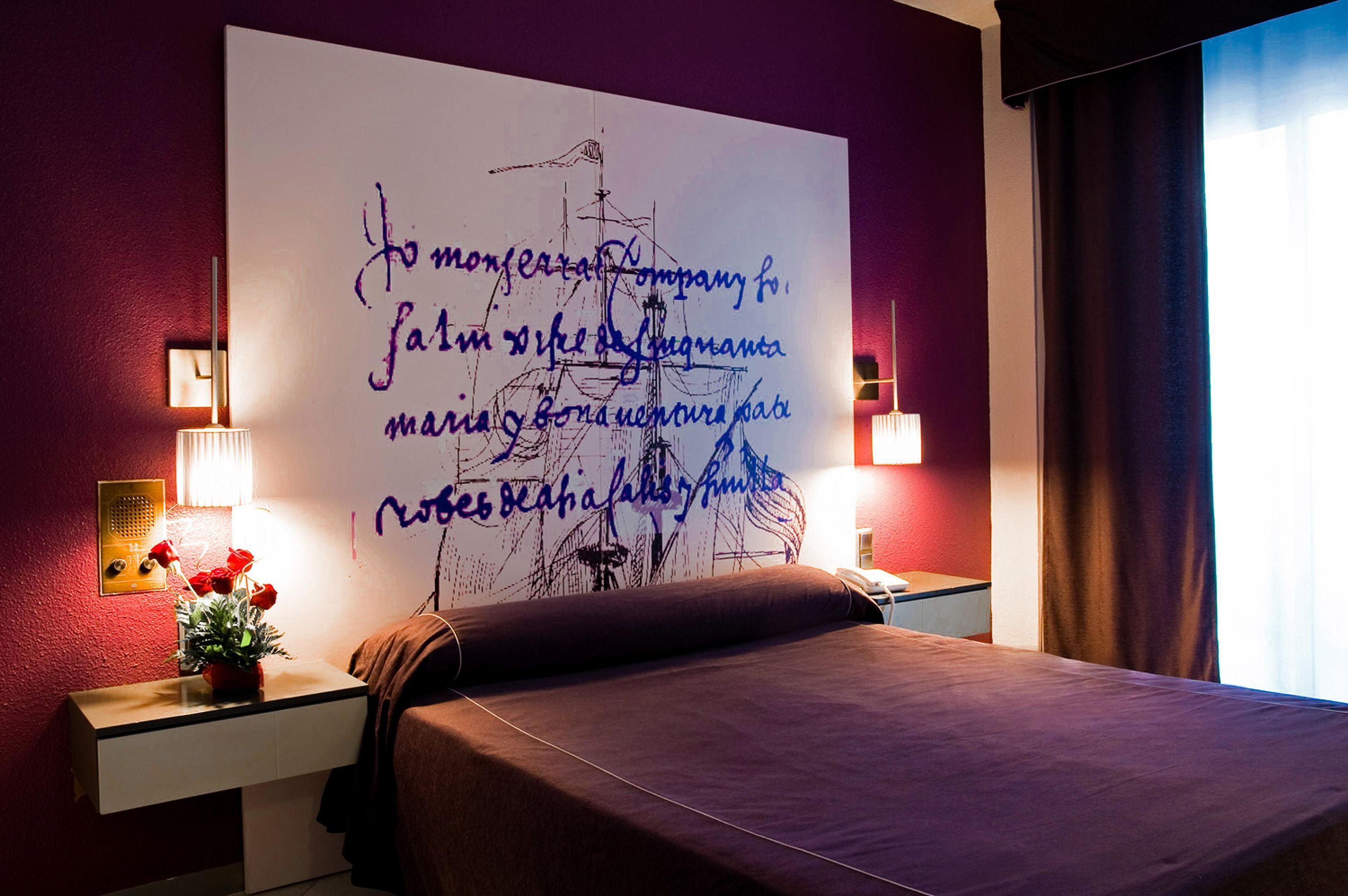 Habitación de hotel con una cama grande, una mesita de noche con lámpara, una pared con un mural decorativo y una ventana con cortinas.