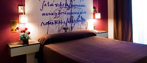 Habitación de hotel con una cama grande, una mesita de noche con lámpara, una pared con un mural decorativo y una ventana con cortinas.
