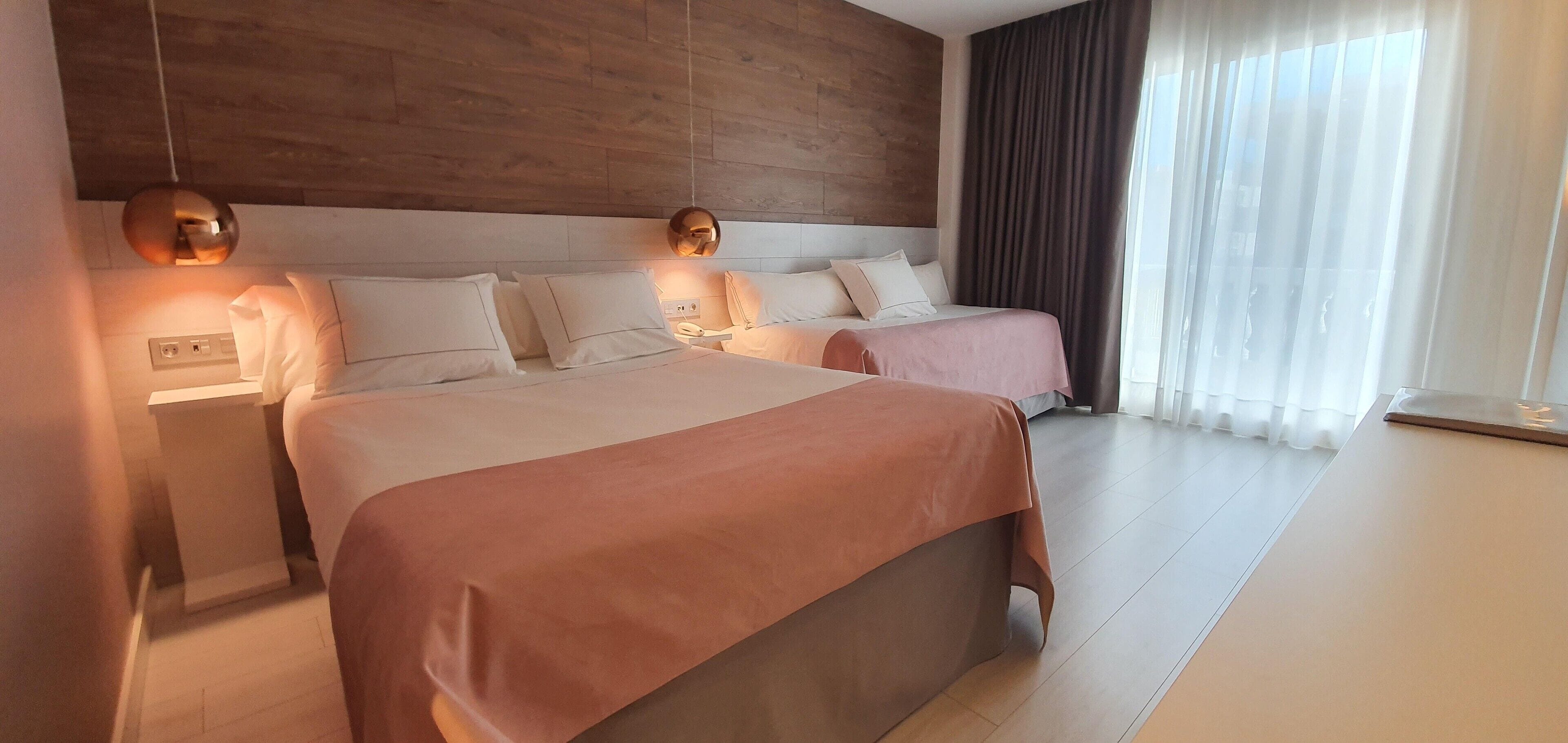 A hotel room with two beds, a wooden headboard, a bedside table with a lamp, and a window with curtains.