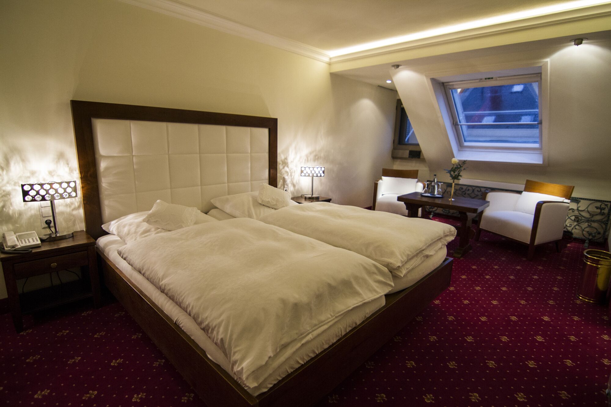A hotel room with a large bed, two bedside tables with lamps, a desk with a chair, and a window with a view.